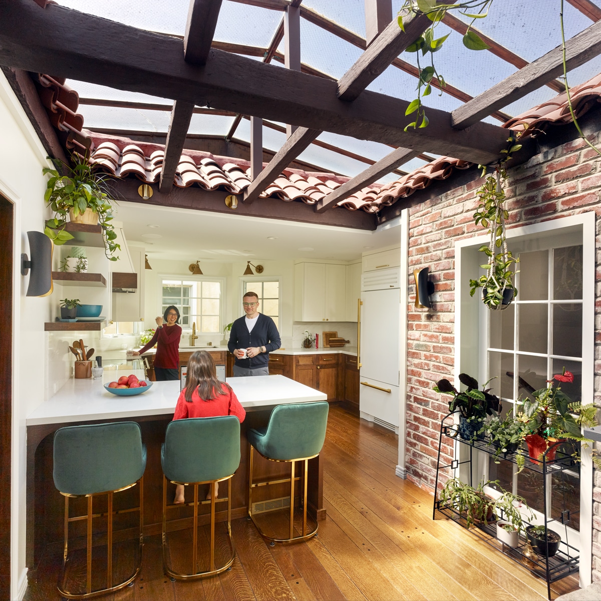 A family of three is in a bright kitchen with a glass ceiling and hanging plants. The parents stand by the island, while a child sits on a green stool. Sunlight streams in, highlighting the wooden floor and brick wall.