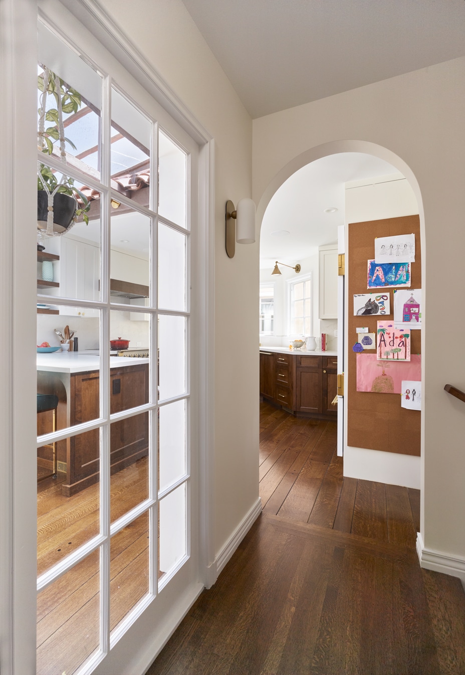 A hallway with wooden floors leads to a kitchen. An archway reveals another room, and a bulletin board on the right wall displays children's colorful artwork. Glass doors on the left open to the kitchen area