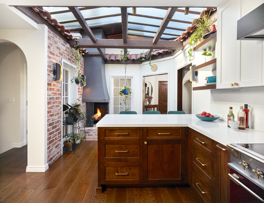A modern kitchen with a large wooden island, white cabinets, and exposed brick walls. Skylights and plants brighten the space, and a lit fireplace is visible in the corner. The floor is hardwood.