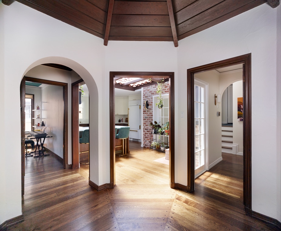 View of a sunlit home interior with wooden floors and ceiling, showing three doorways leading to a dining area, a kitchen with hanging plants, and a hallway with stairs.