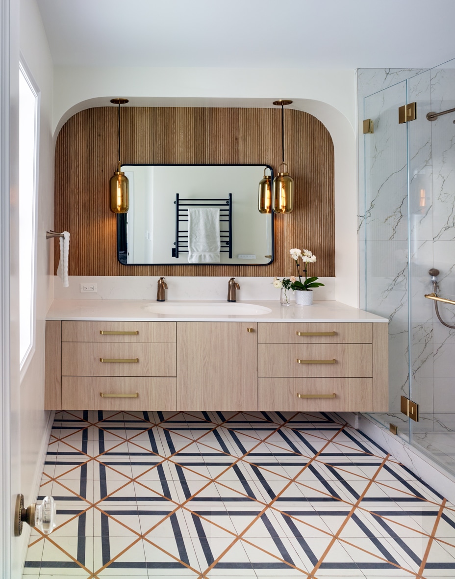 Modern bathroom with a geometric tiled floor, light wood vanity, brass fixtures, marble shower, large mirror with wooden paneling, and two hanging brass pendant lights. A white towel and a small orchid plant add accents