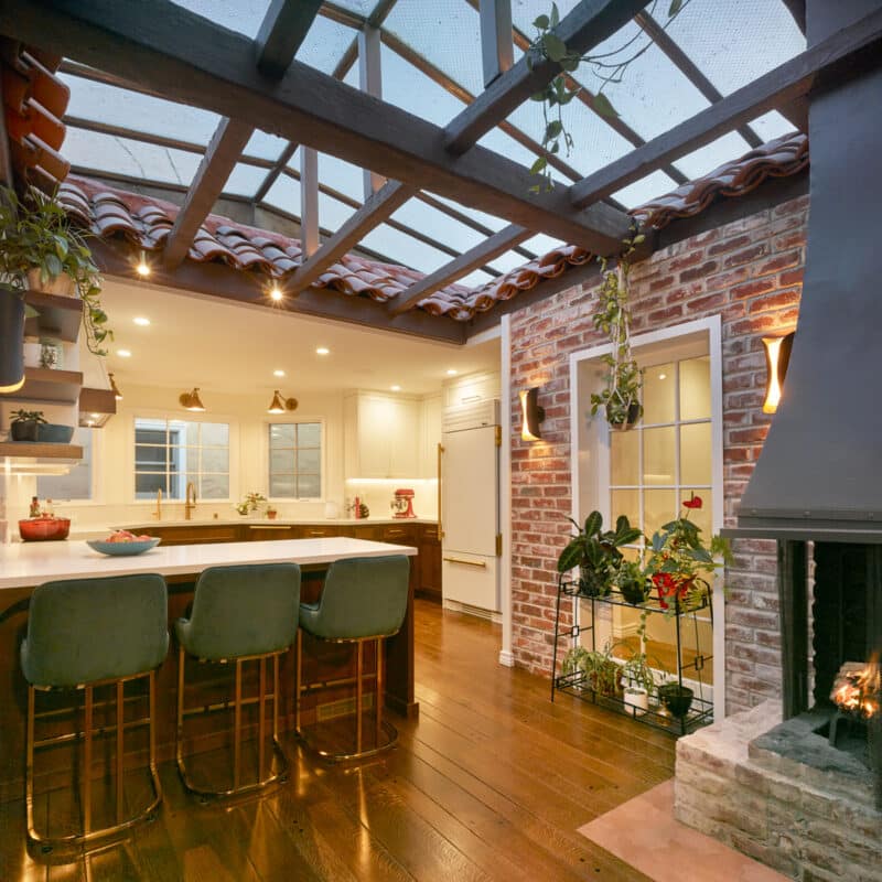 A cozy kitchen with a skylight roof, exposed brick walls, a fireplace, green cushioned bar stools at a white island, hanging plants, and wooden floors, warmly lit with recessed lighting.