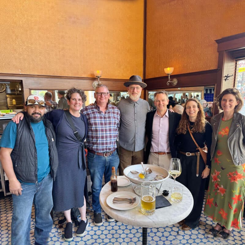 Seven people stand smiling together inside a restaurant, gathered around a small round table with drinks and plates. The setting has warm-colored walls, large windows, and tiled floors.