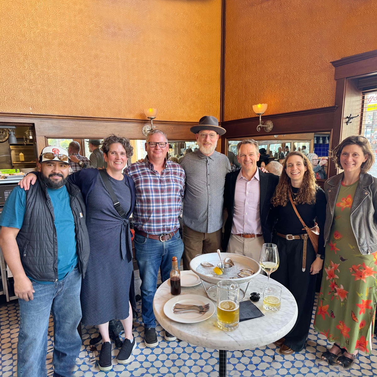 Seven people stand smiling together inside a restaurant, gathered around a small round table with drinks and plates. The setting has warm-colored walls, large windows, and tiled floors.