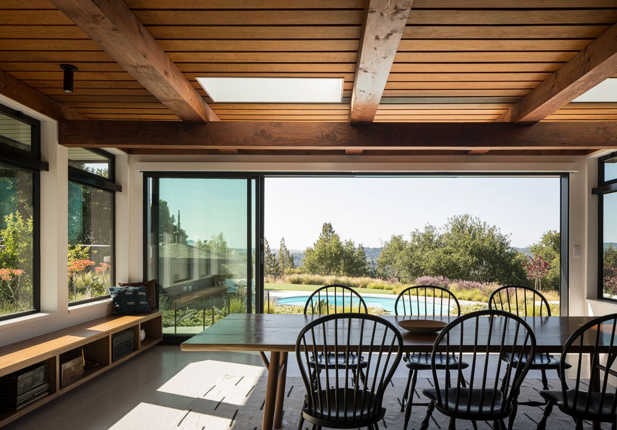 A modern dining room with wooden ceiling beams, black chairs around a long table, large sliding glass doors, and a view of a garden, pool, and trees outside. Sunlight fills the space, creating a warm atmosphere.