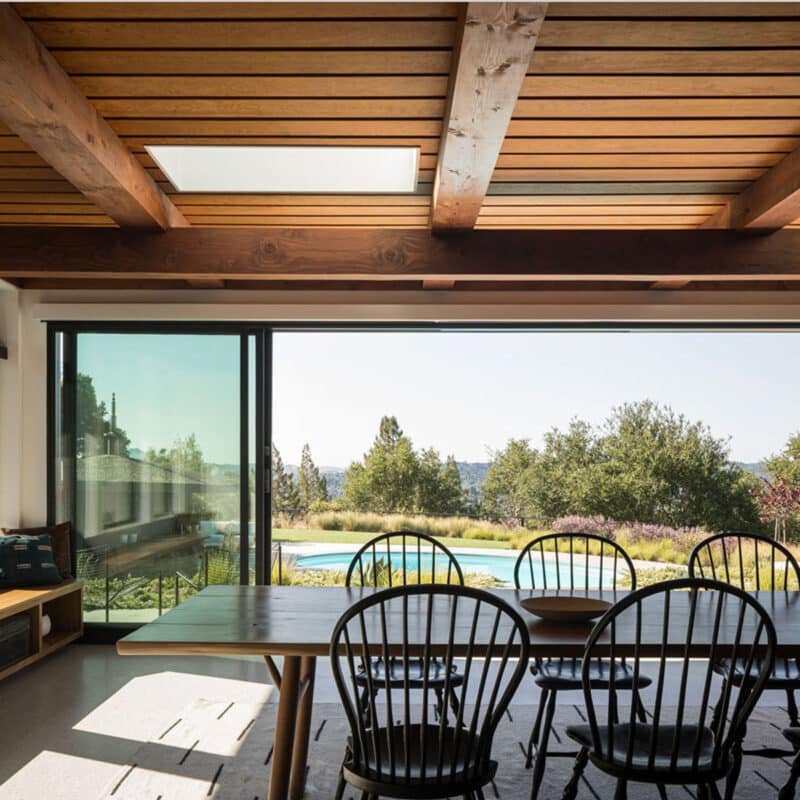 A modern dining room with wooden ceiling beams, black chairs around a long table, large sliding glass doors, and a view of a garden, pool, and trees outside. Sunlight fills the space, creating a warm atmosphere.