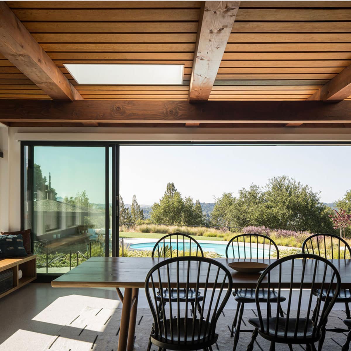A modern dining room with wooden ceiling beams, black chairs around a long table, large sliding glass doors, and a view of a garden, pool, and trees outside. Sunlight fills the space, creating a warm atmosphere.