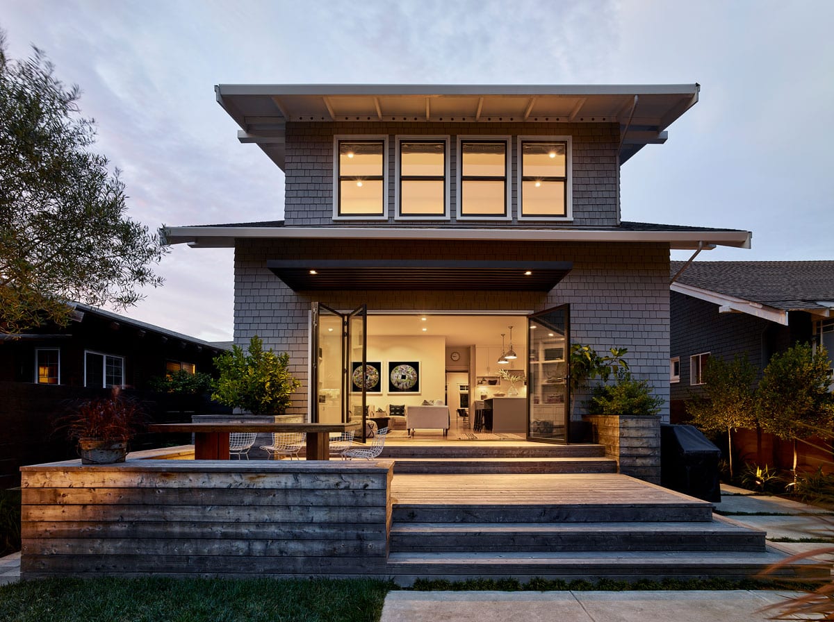 Modern two-story house with gray shingles, large windows, and an open ground floor leading to a wooden deck with built-in seating, outdoor plants, and evening interior lights visible