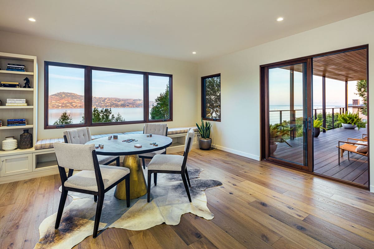 Modern dining area with a round table and four chairs on a cowhide rug, large windows offering a scenic water view, built-in shelves, wooden floors, and a sliding glass door leading to a furnished balcony with plants.