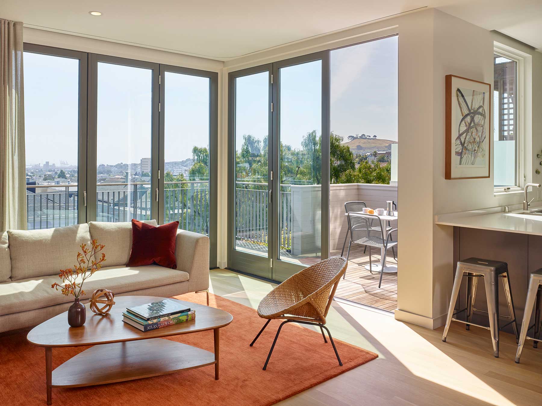 A bright, modern living room with floor-to-ceiling windows and a balcony. Features a beige sofa, a wooden coffee table, a wicker chair, an orange rug, and a kitchen island with stools. Sunlight fills the space.