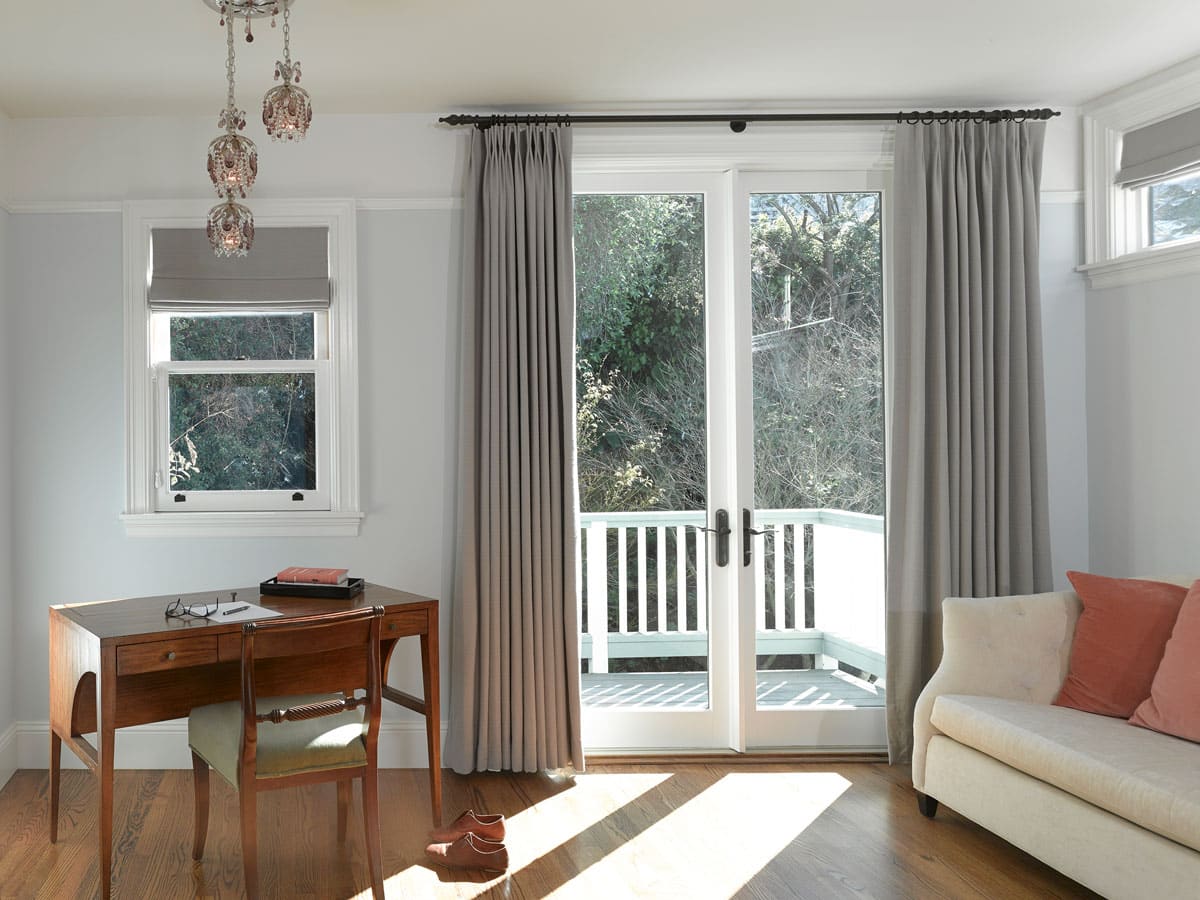 A bright room with a wooden desk and chair, an open notebook, glasses, and a pen on the desk, a sofa with pillows, and large glass doors with gray curtains leading to a balcony with trees outside.